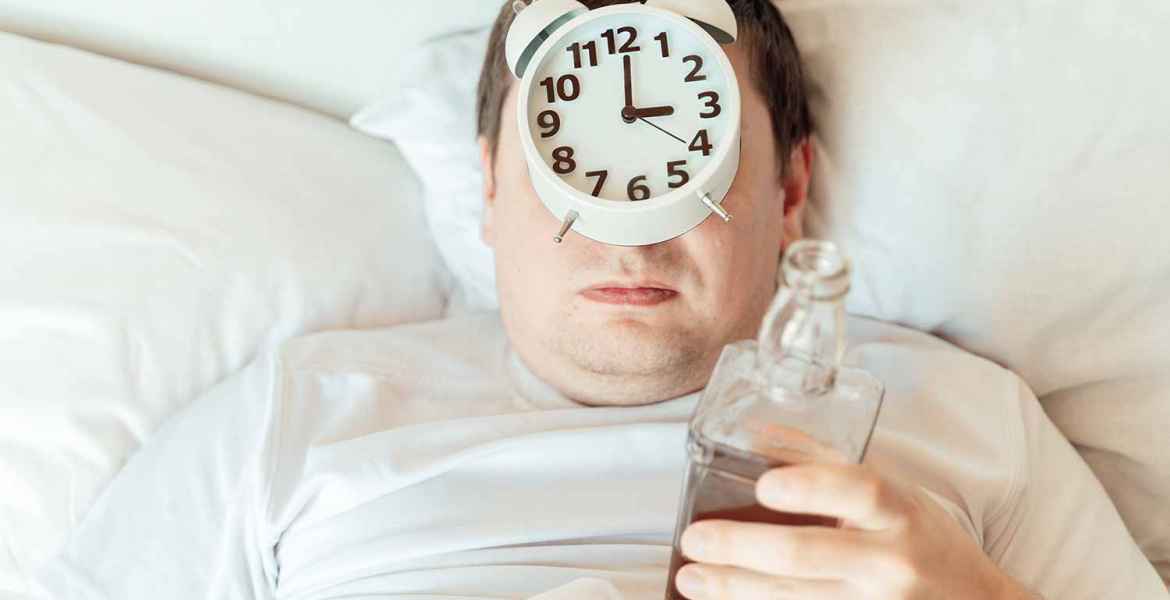 Man can't sleep at night with half empty bottle and alarm clock on his face. Insomnia after drinking alcohol.