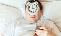 Man can't sleep at night with half empty bottle and alarm clock on his face. Insomnia after drinking alcohol.