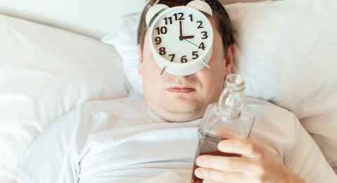 Man can't sleep at night with half empty bottle and alarm clock on his face. Insomnia after drinking alcohol.