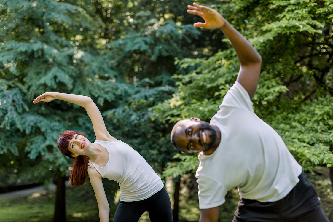 active healthy couple, african guy and caucasian girl, working out together outdoors, doing stretching exercises on the grass. sport and fitness concept. focus on woman behind